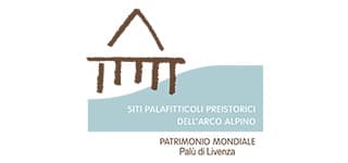 Logo del Patrimonio Mondiale di Palù di Livenza, con il disegno marrone di una palafitta stilizzata sopra un riquadro azzurro. Sotto, la scritta Siti palafitticoli preistorici dell’arco alpino e Patrimonio Mondiale Palù di Livenza.