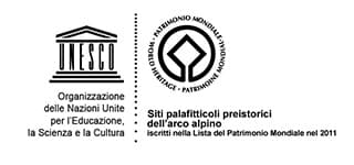 Il logo UNESCO, raffigurato come un tempio, e la scritta Organizzazione delle Nazioni Unite per l’Educazione, la Scienza e la Cultura. Accanto, il simbolo circolare del Patrimonio Mondiale con un rombo al centro e le scritte Siti palafitticoli preistorici dell’arco alpino Iscritti nella Lista del Patrimonio Mondiale nel 2011.