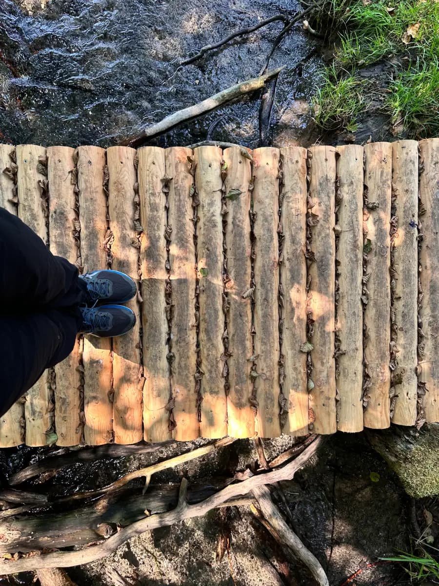 Una persona con pantaloni neri e scarpe da trekking blu si trova su un ponte di assi formato da assi di legno affiancate.